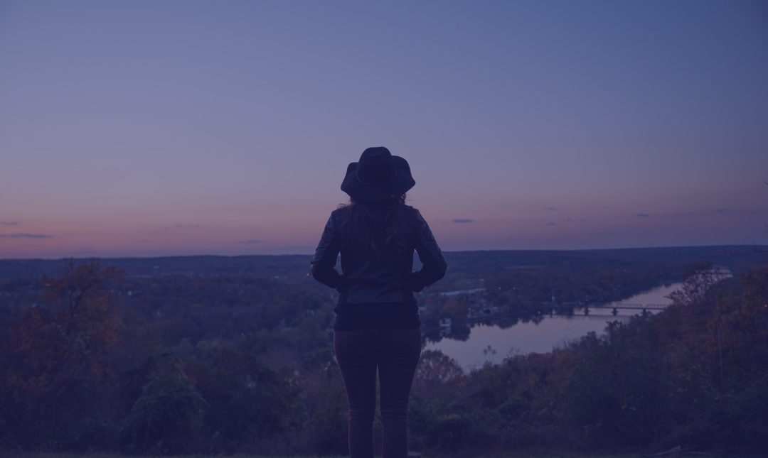 A woman looking over the river from a high hill at dusk.